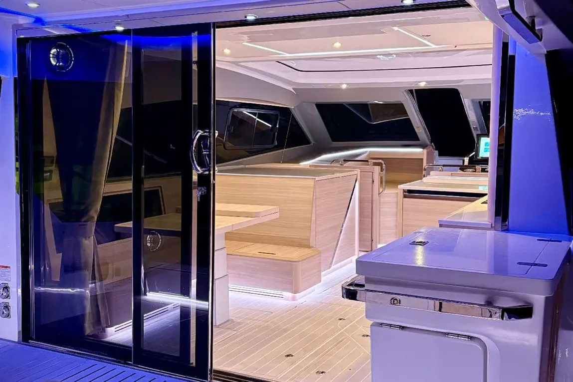 Sleek interior of a modern yacht featuring a glass door, wooden furniture, and ambient lighting. A kitchen area is visible.